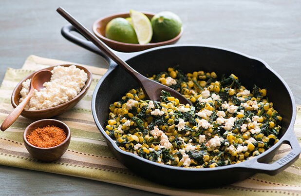 A skillet with corn, spinach and cotija cheese on a napkin beside bowls of cheese and cayenne.